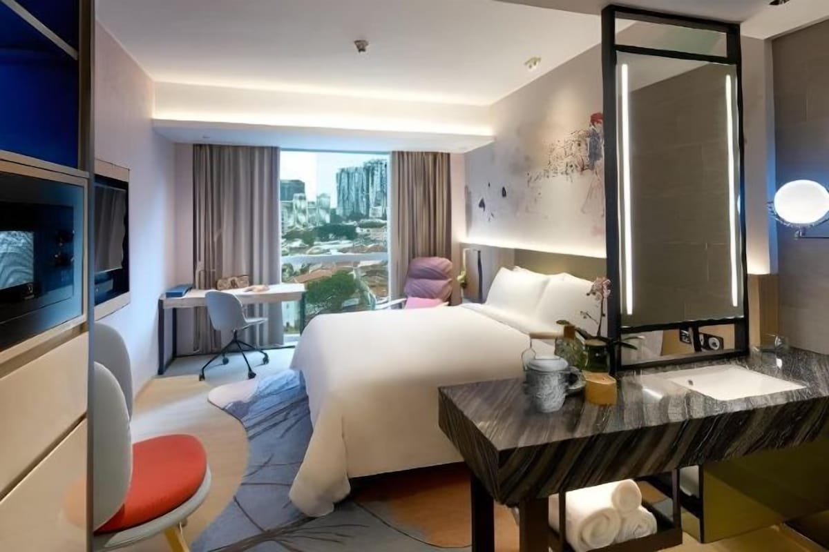 A hotel room with a bed, a desk, a chair, a large mirror, and a view of the cityscape.