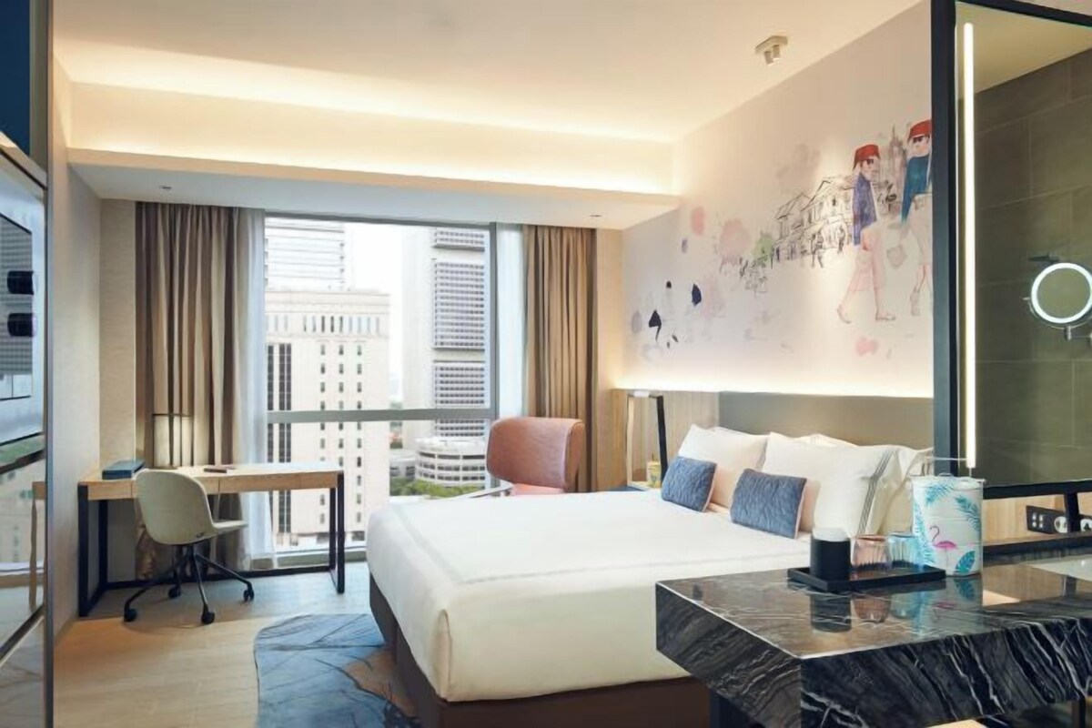 A modern hotel room with a large bed, a desk, and a computer. There is a large window with a view of buildings outside.