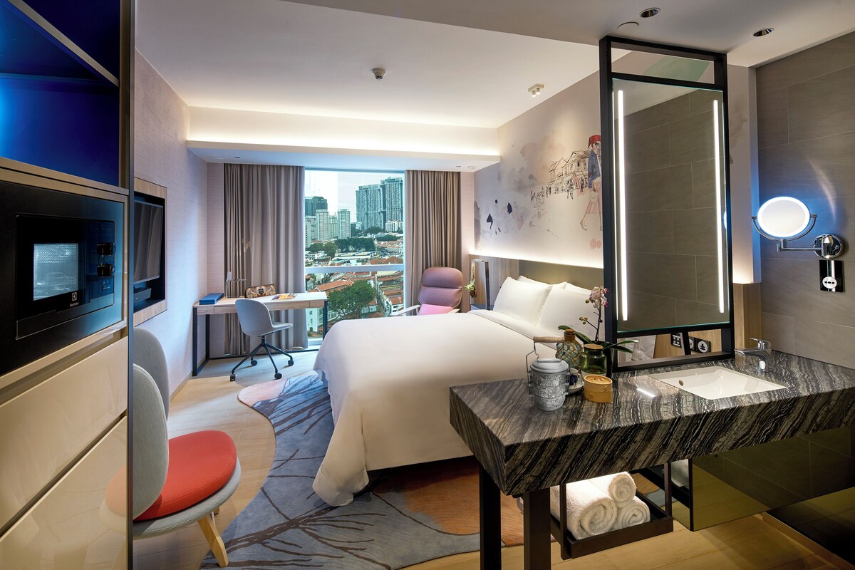 A hotel room with a bed, desk, chair, TV, and a view of the city.