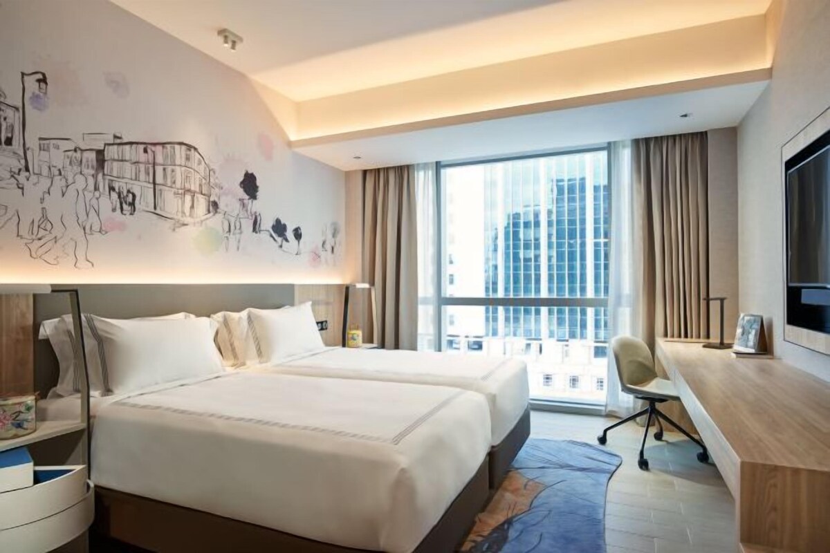 A hotel room with a large bed, a desk, a chair, and a large window with a city view.
