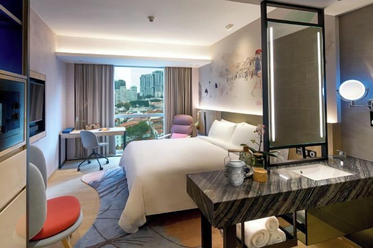 A hotel room with a large bed, a desk, a chair, a TV, a bathroom with a mirror and sink, and a view of the city.