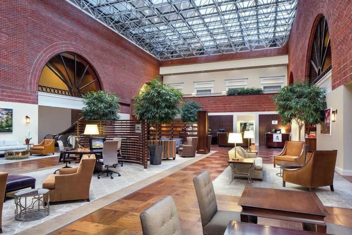 A spacious lobby with a high ceiling, brick walls, and a central seating area with tables and chairs.