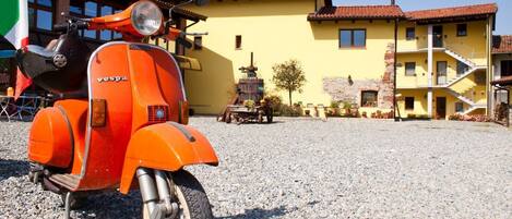 A vintage orange Vespa scooter parked in front of a yellow building with a stone facade and a red-tiled roof.