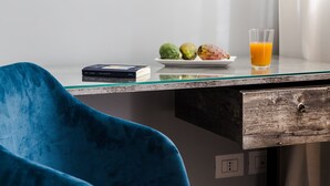 Una habitación moderna con un escritorio de vidrio, una silla azul, un libro, un plato con frutas y un vaso de zumo de naranja.