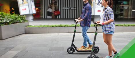A man and a woman on electric scooters in an urban setting with a modern building in the background.