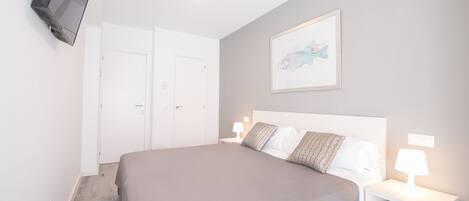 A bedroom with a large bed, white walls, a mounted TV, a framed picture, and bedside lamps.