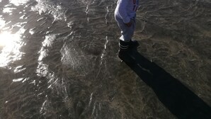 Un bambino gioca in acque basse su una spiaggia, con barche e un molo roccioso sullo sfondo.