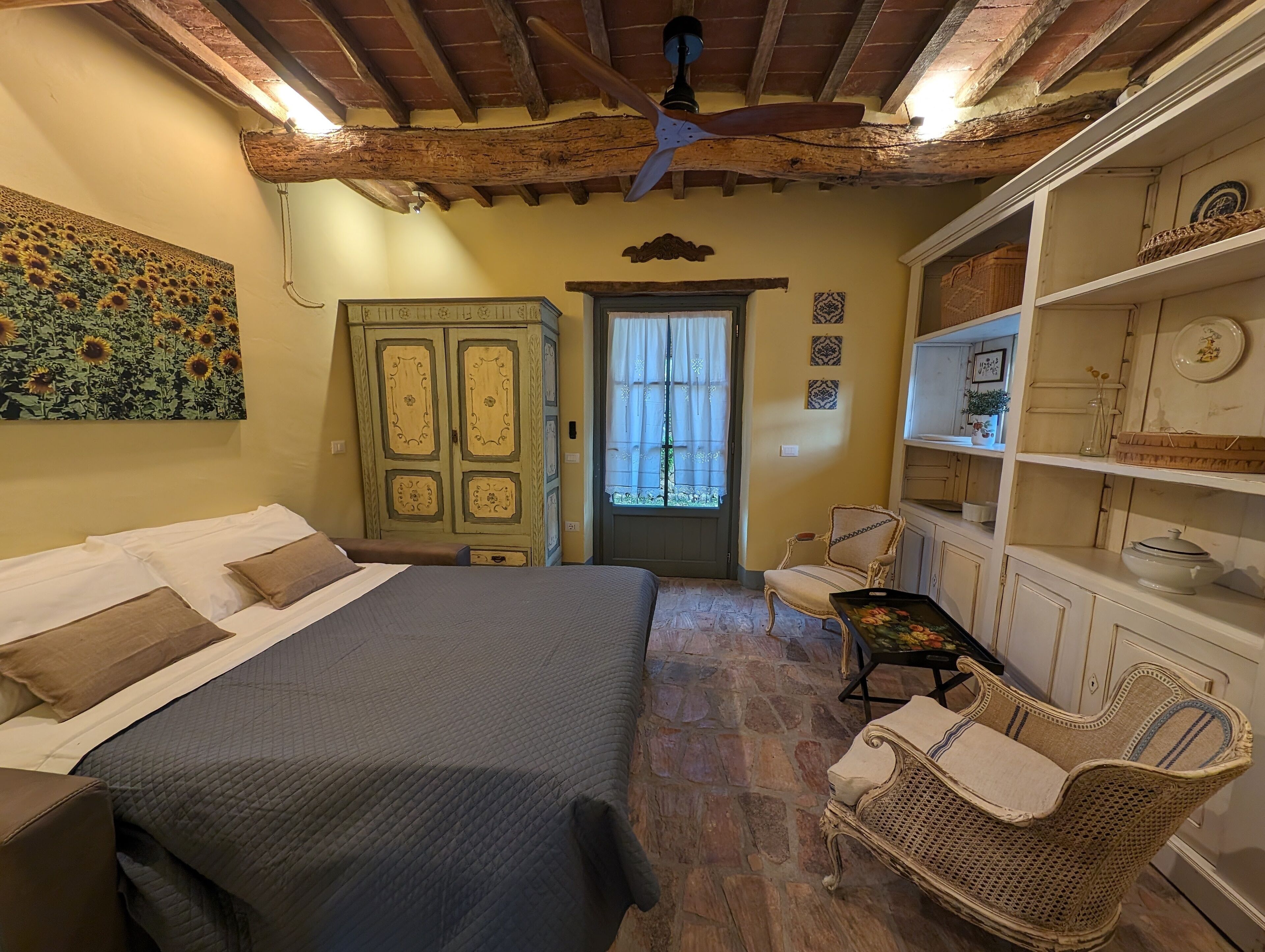 Una habitación con cama, una silla, estante con adornos, puerta con ventana y ventilador de techo.