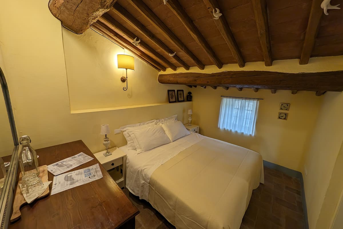 A bedroom with a wooden ceiling, a bed with white bedding, a nightstand with a lamp, and a wooden dresser with a mirror and glassware.