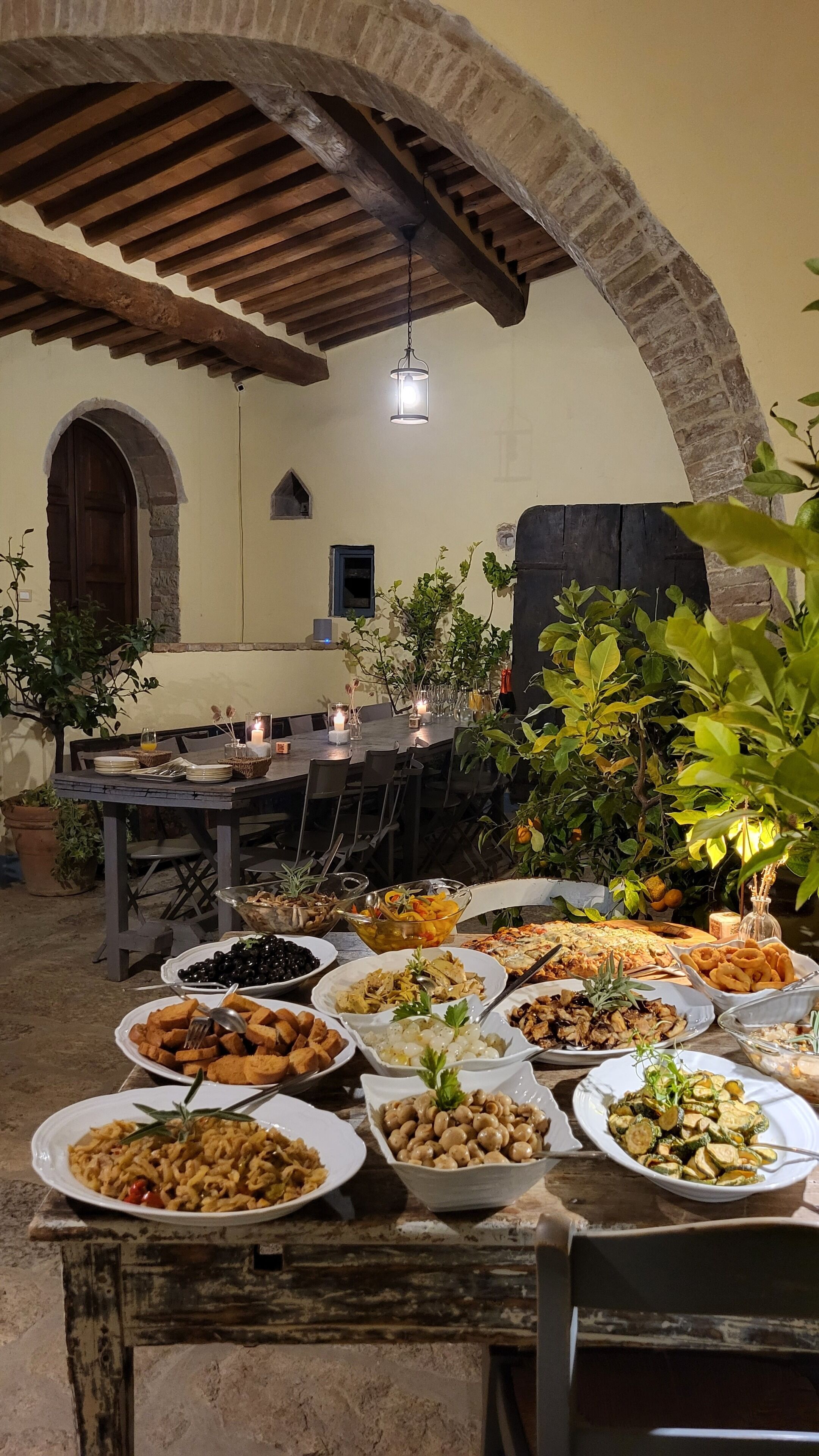 Un espacio exterior para comer con un arco de piedra rústica, vigas de madera y una variedad de platos sobre una mesa larga.