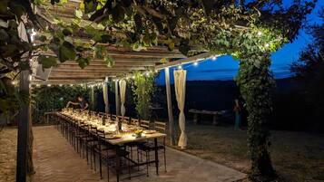 Un área de comedor al aire libre con una mesa larga lista para una comida, bajo una pérgola adornada con luces colgantes.