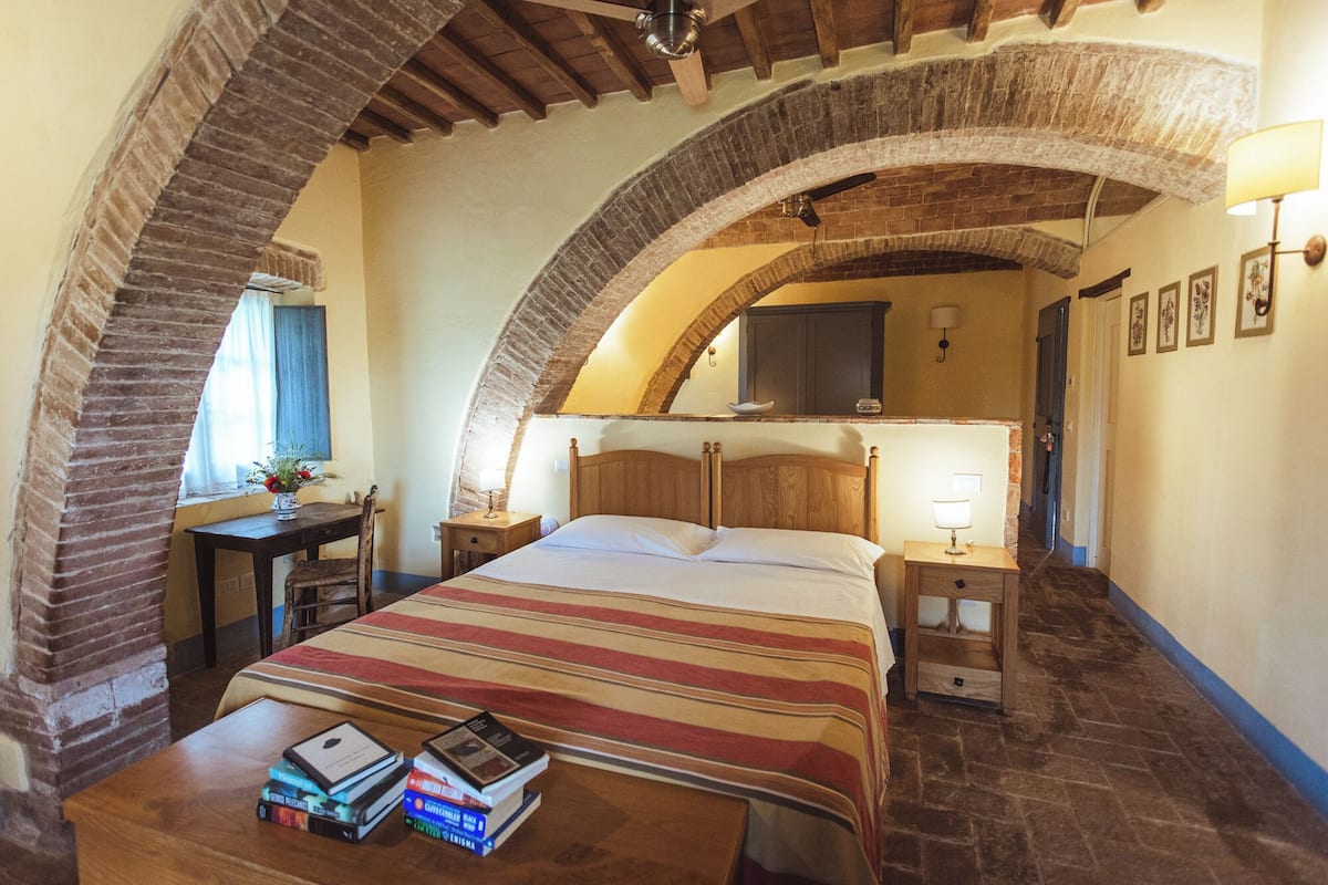 A bedroom with a wooden bed, a bedside table with books, a lamp, and a view of a bathroom through an arched brick ceiling.