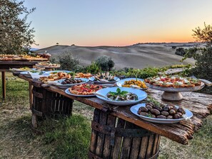 Un espace de restauration en plein air, avec différentes assiettes disposées sur des tables en bois rustique, le tout mis en valeur par un paysage pittoresque de collines ondulantes et un coucher de soleil.