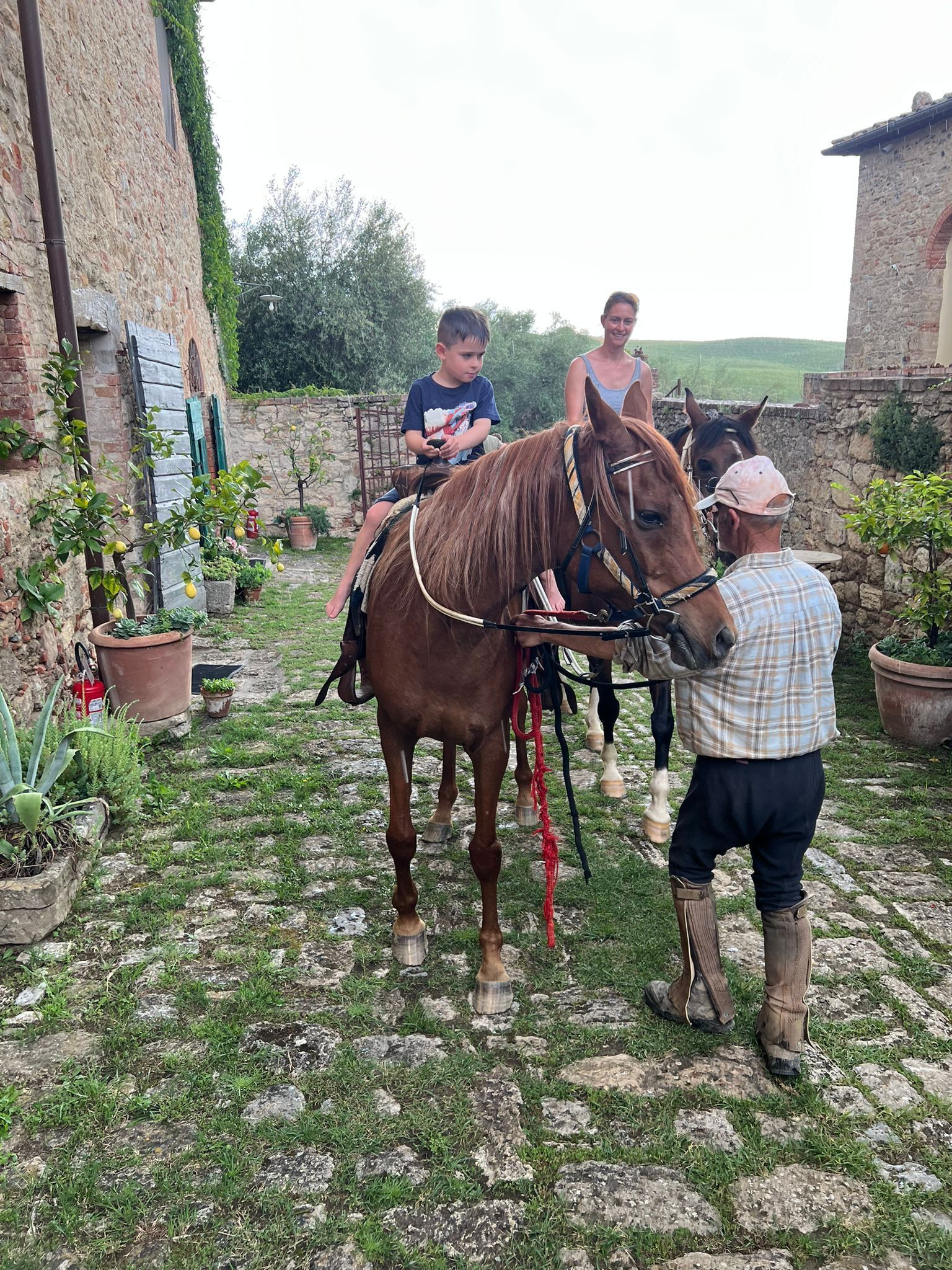 Un cheval, avec son cavalier et son accompagnateur, dans un décor champêtre en plein air, agrémenté de murs en pierre et de plantes en pot.
