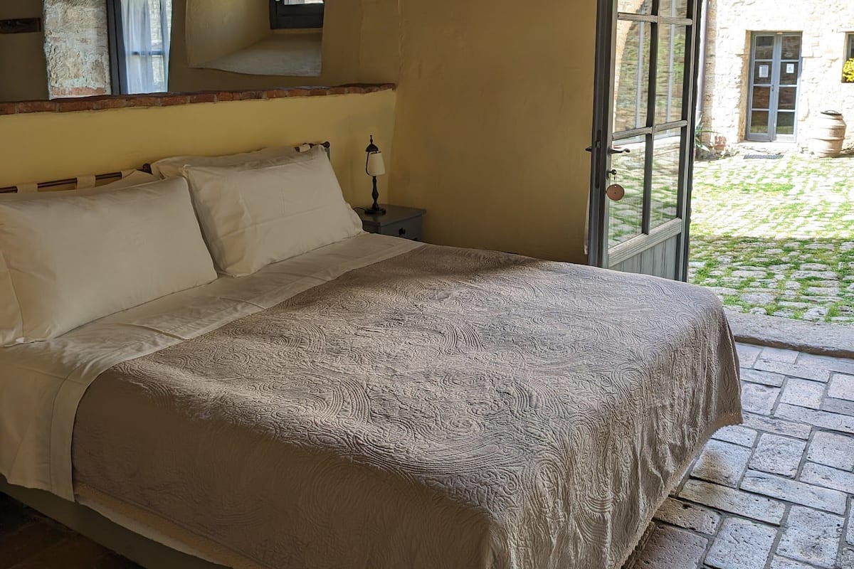 A bedroom with a bed, a nightstand, a lamp, and a window with a view of the outside.