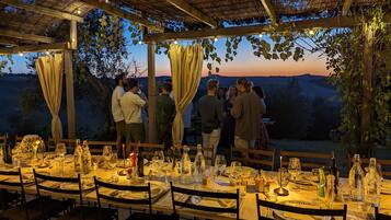 An outdoor dining area with tables set for a meal, surrounded by a rustic pergola with string lights.