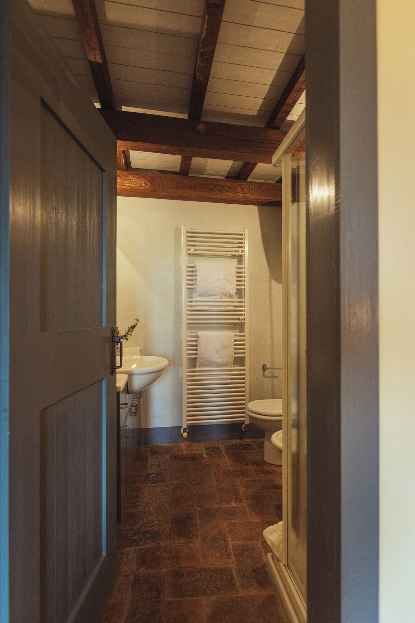 A narrow hallway with a wooden door on the left, leading to a bathroom with a white sink, toilet, and towel rack.
