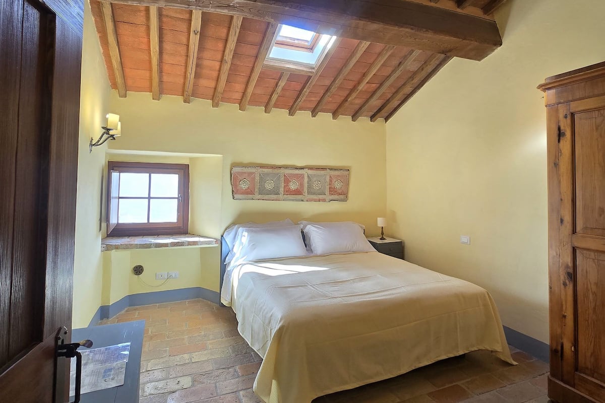 A bedroom with a wooden ceiling, a skylight, a bed with a beige bedspread, a nightstand, a wall clock, and a wooden wardrobe.