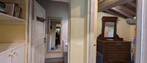 A narrow hallway with a wooden dresser and a mirror. A door leads to another room.