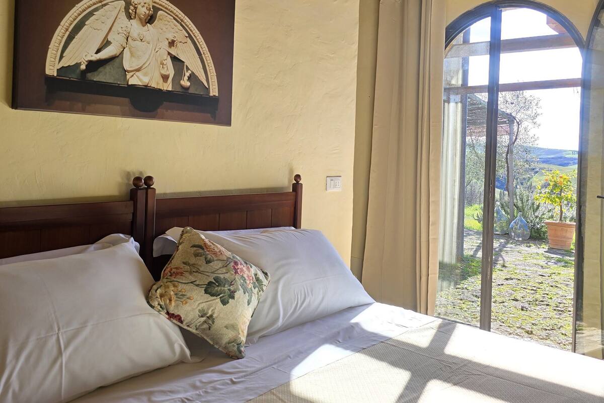 A bed with white linens and a floral pillow, a wooden headboard, a wall-mounted light fixture, and a view of the outdoors through a window with a curtain.