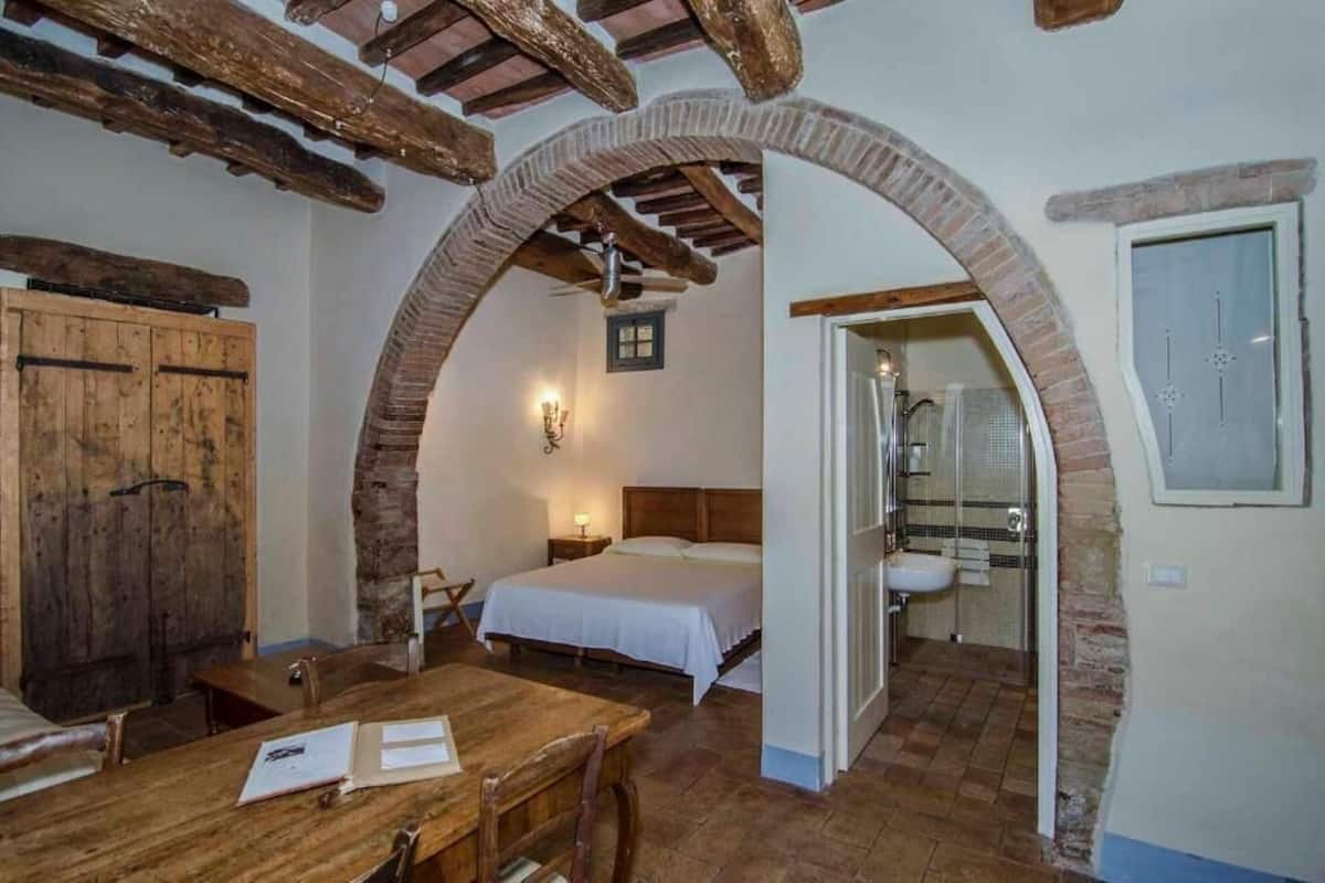 A rustic room with a wooden table, a bed, and a bathroom visible through an archway.