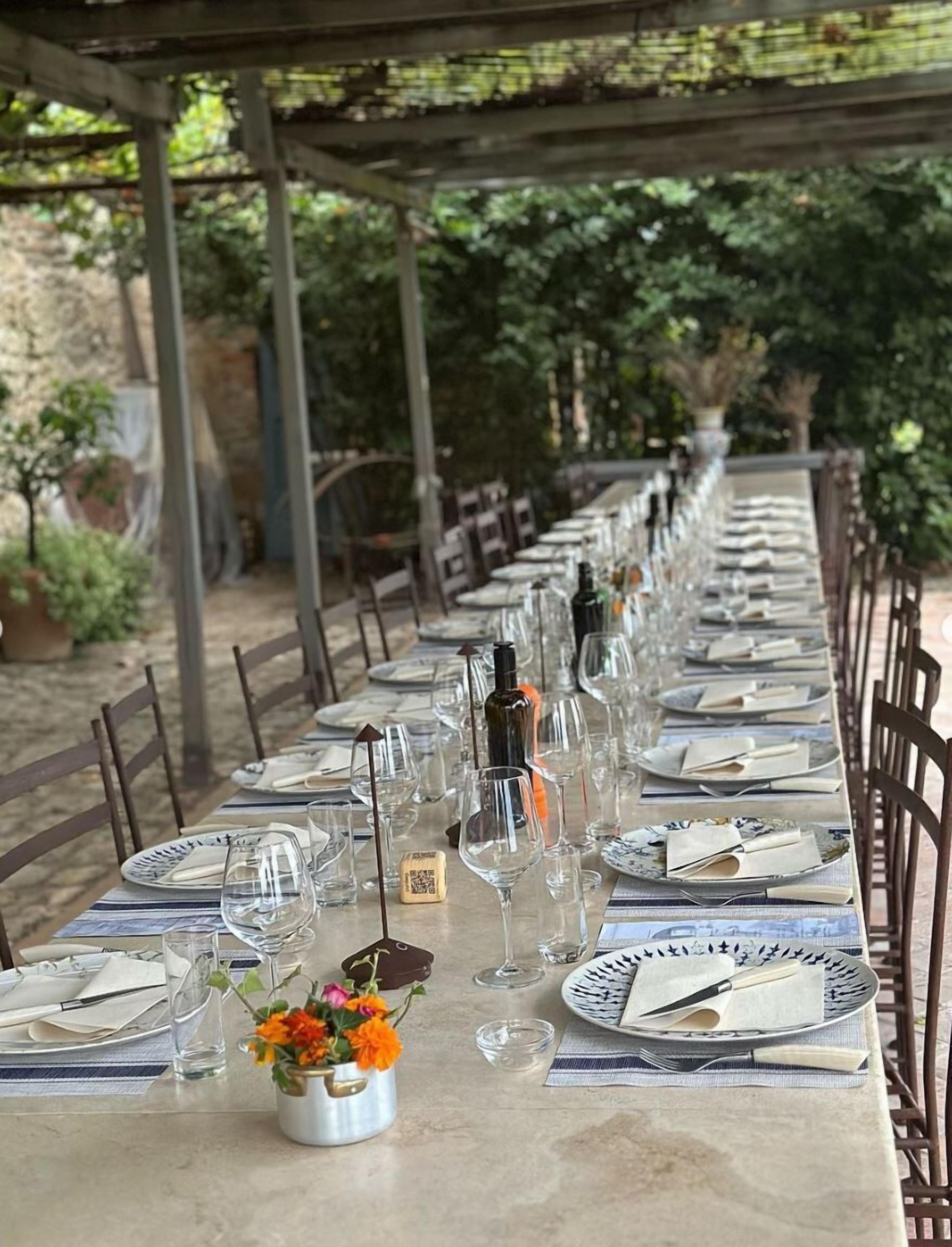 A long outdoor dining table set with plates, glasses, and cutlery, surrounded by chairs.