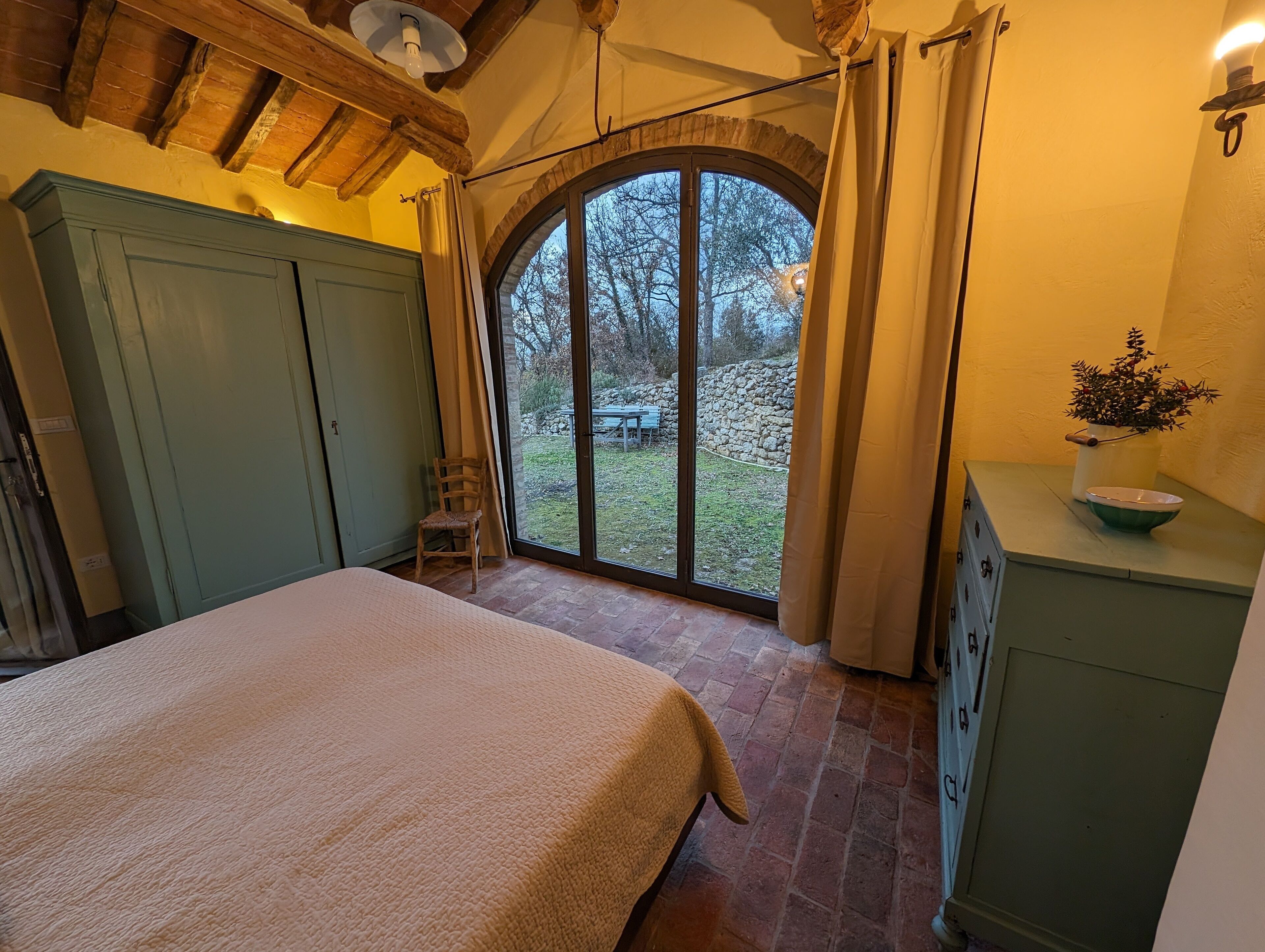 Een slaapkamer met een groot bed, een stoel, een houten garderobe, een klein tafeltje met een schaal en uitzicht op de omgeving via een glazen deur.