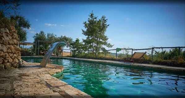 Una piscina con pared de piedra y escalera de metal, rodeada de árboles y un cielo despejado.