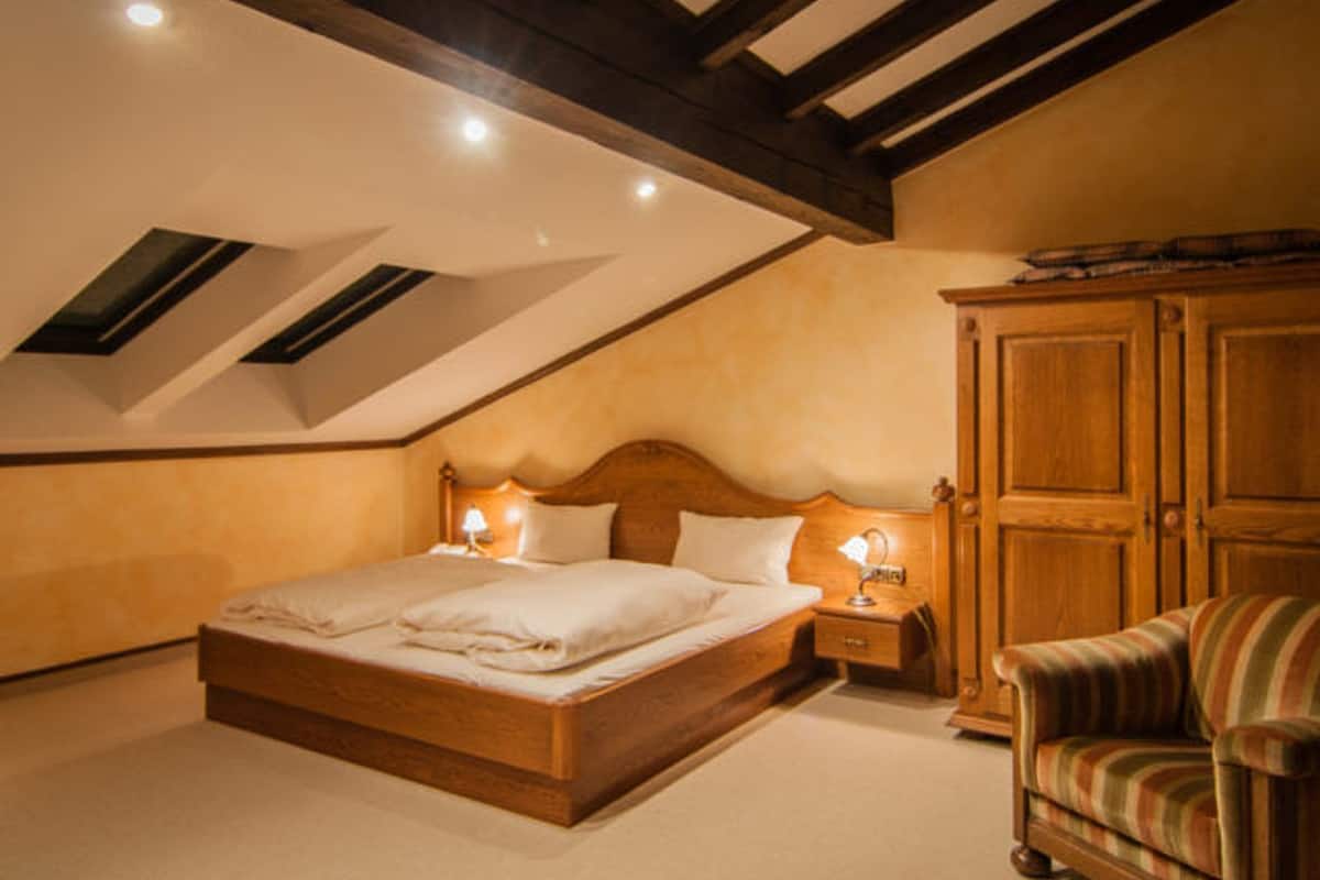 A bedroom with a sloped ceiling, a wooden bed, bedside lamps, a wardrobe, and a striped armchair.