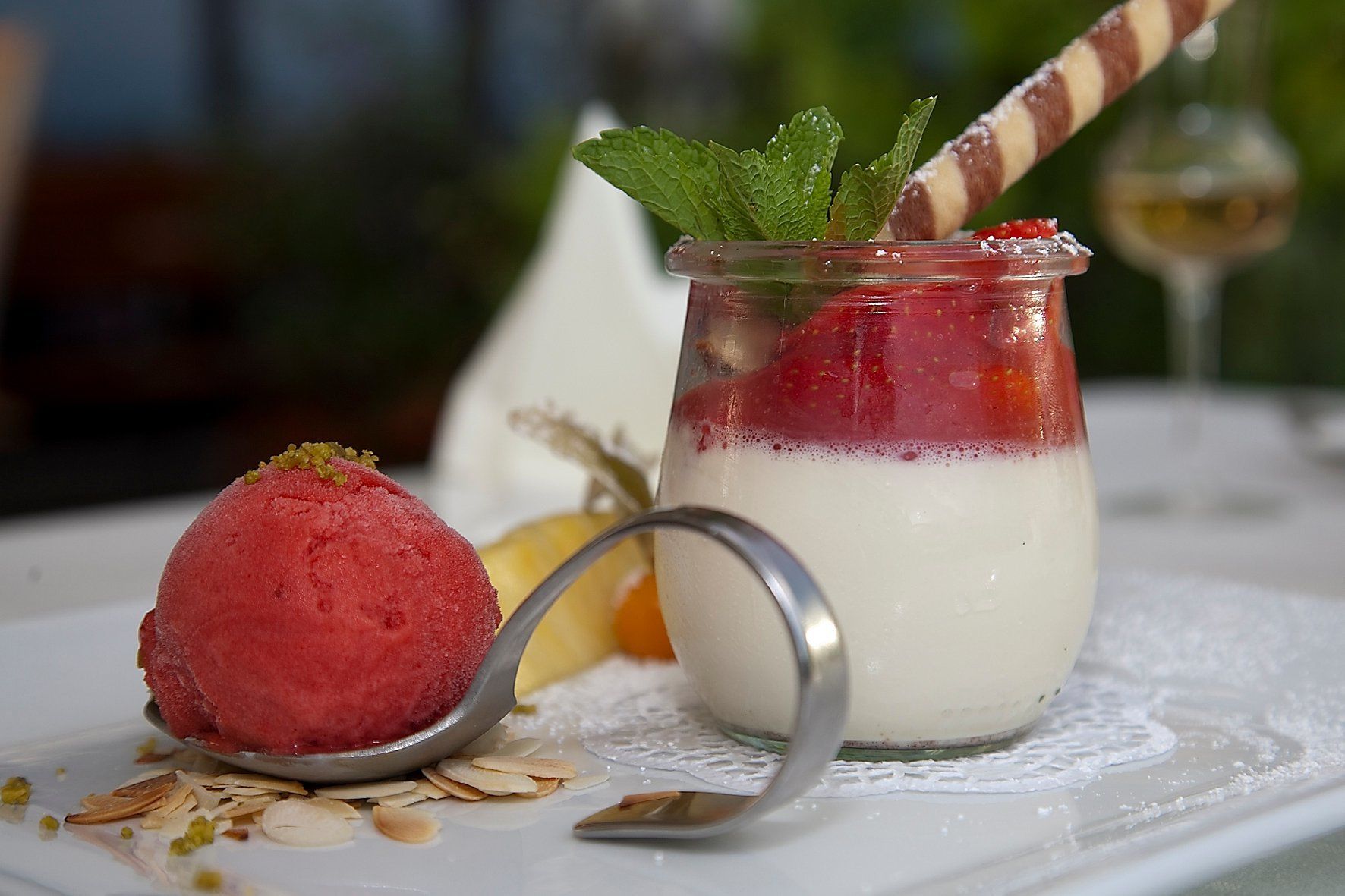 En dessert med en skopa jordgubbssorbet och en drink i ett glas, garnerad med mynta och mandlar, serverad på ett vitt bordsduk.