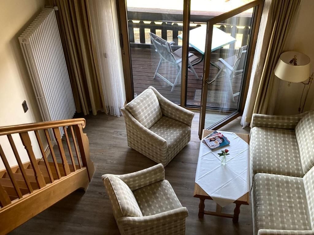A room with a wooden staircase, a checkered sofa, a table with a white tablecloth, and a view of an outdoor deck with chairs.