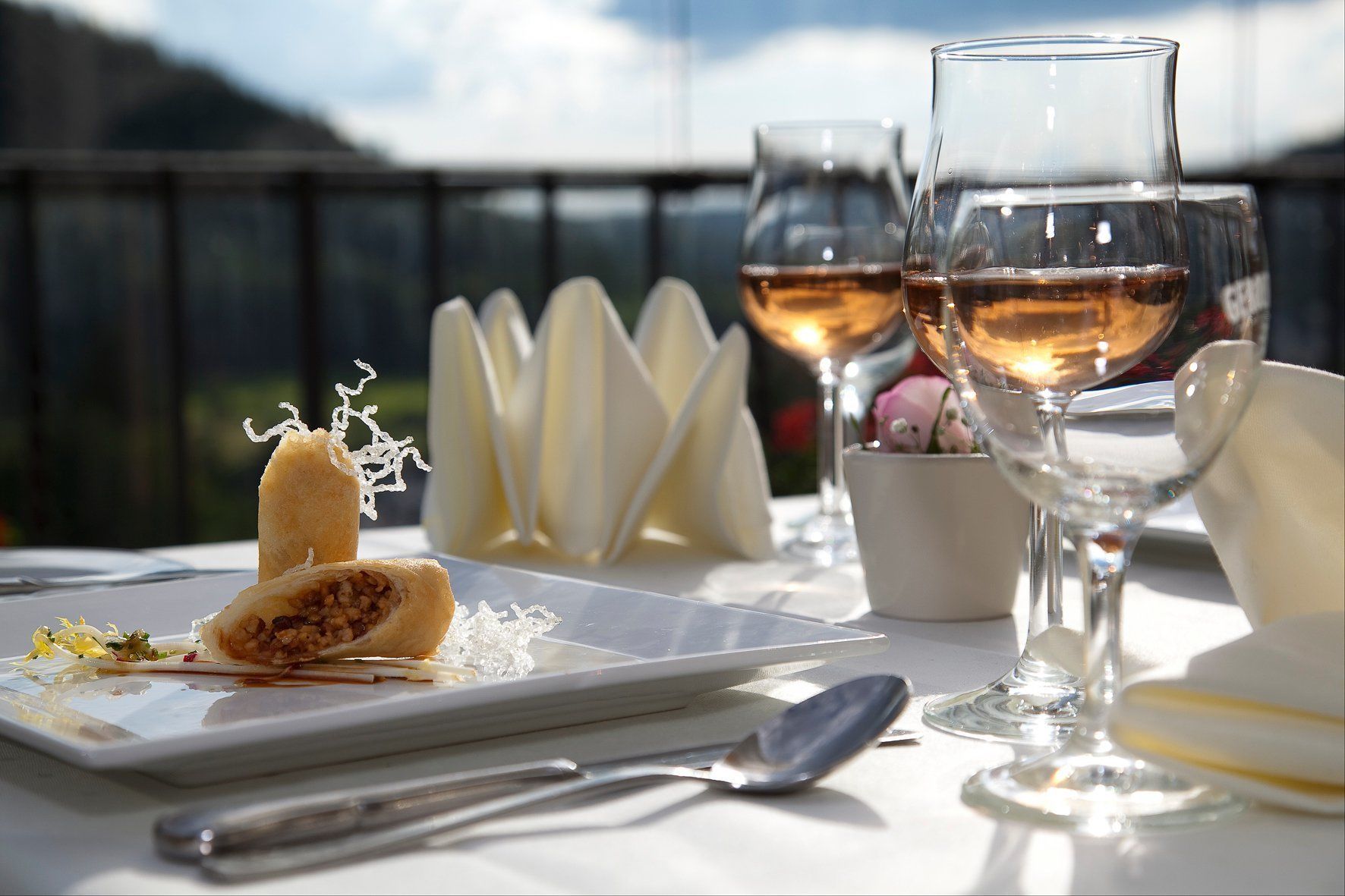 A table with a dessert, two wine glasses, and a spoon on a terrace with a view.