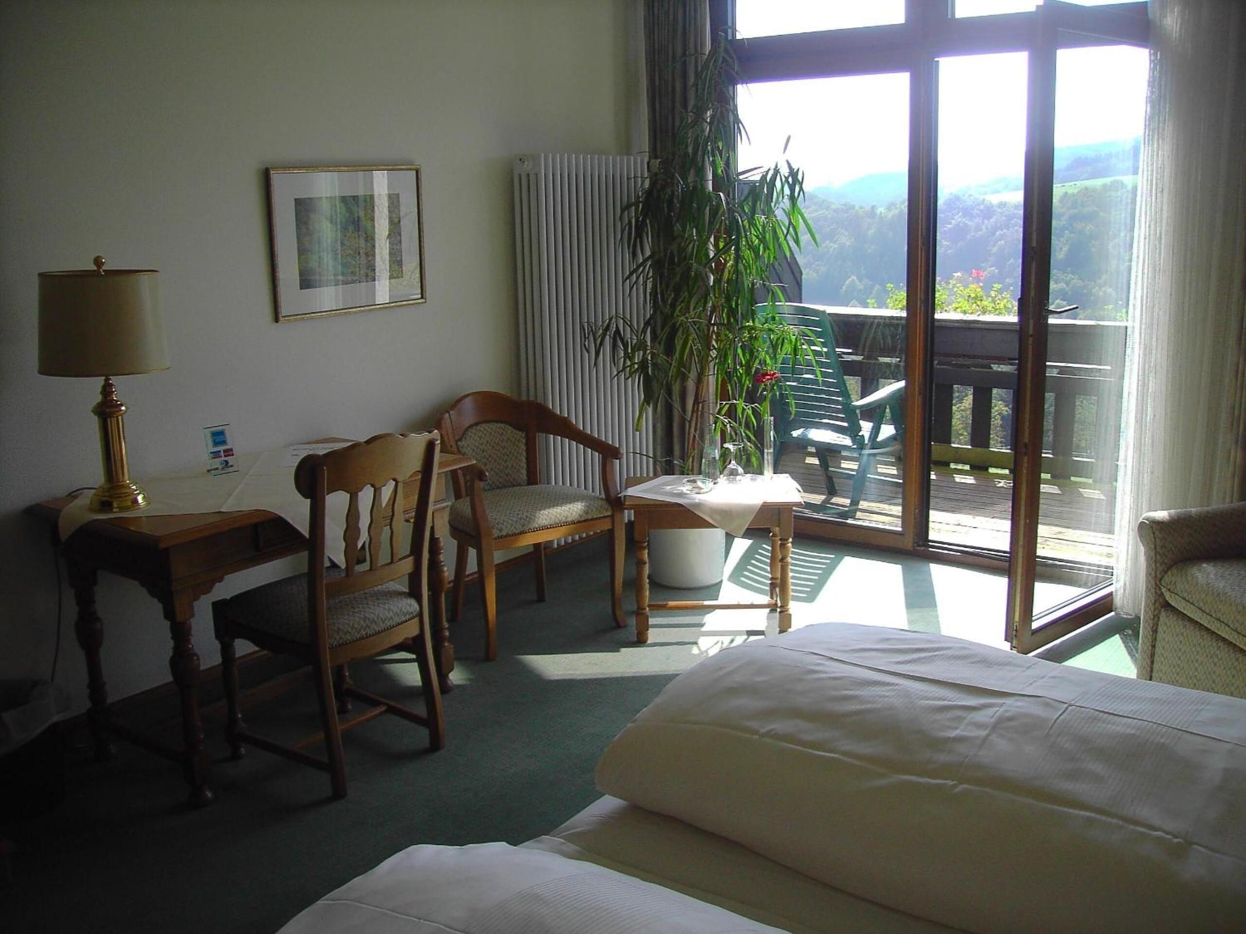 Ein Hotelzimmer mit einem Bett, einem Schreibtisch mit Lampe, zwei Stühlen, einem Tisch mit Vase und einem großen Fenster mit Blick ins Freie.