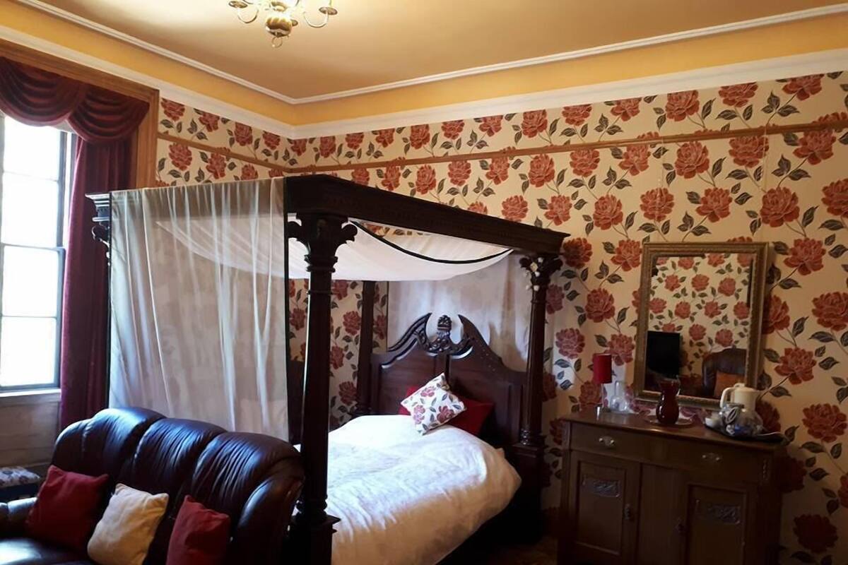 A four-poster canopy bed with a wooden headboard, a leather sofa with cushions, a wooden nightstand with a mirror, and floral wallpaper.