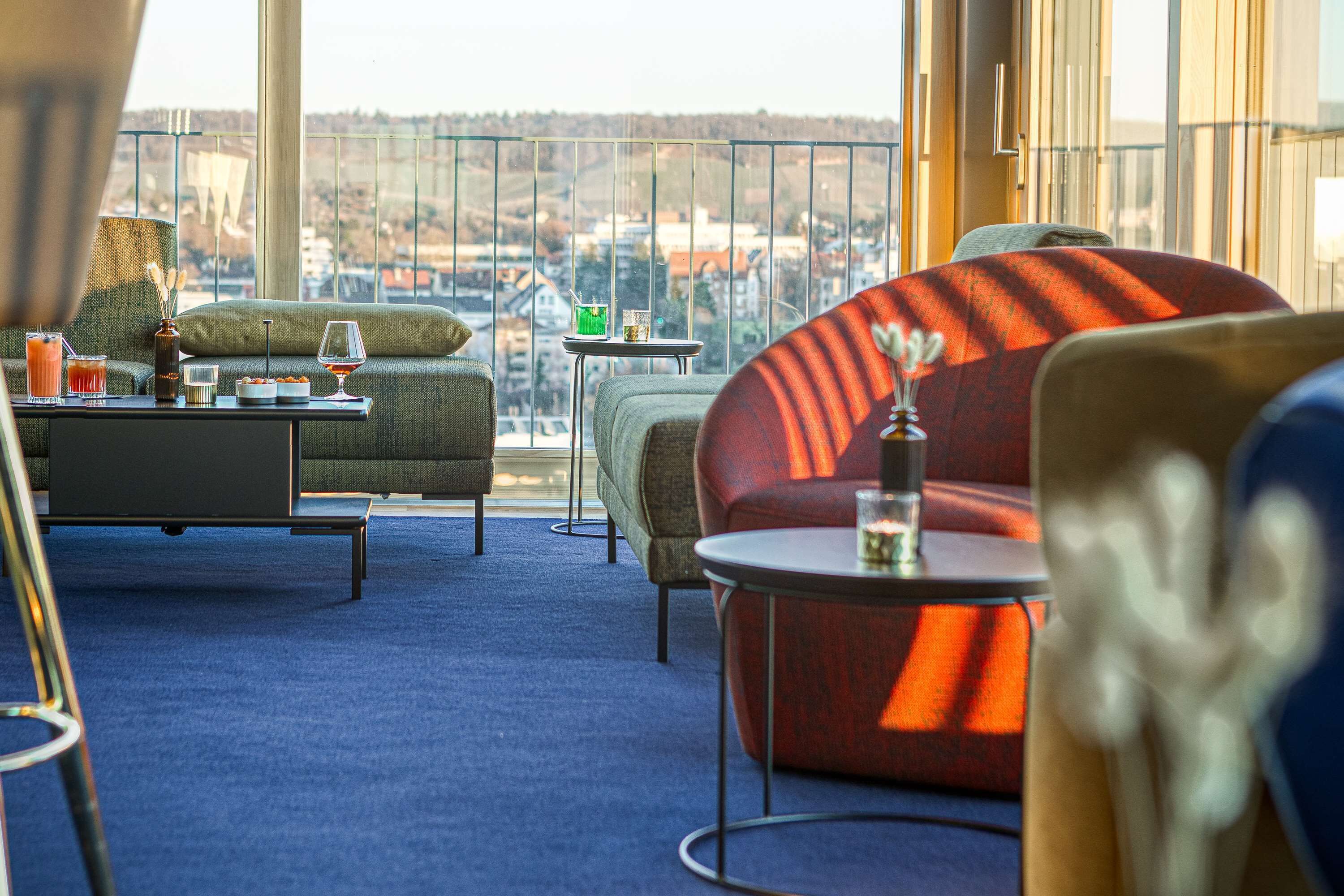 Uma área de lounge moderna com vista para a paisagem urbana, com um sofá, poltrona e pequenas mesas com bebidas e petiscos.