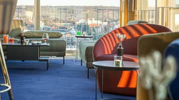 Uma área de lounge moderna com vista para a paisagem urbana, com um sofá, poltrona e pequenas mesas com bebidas e petiscos.