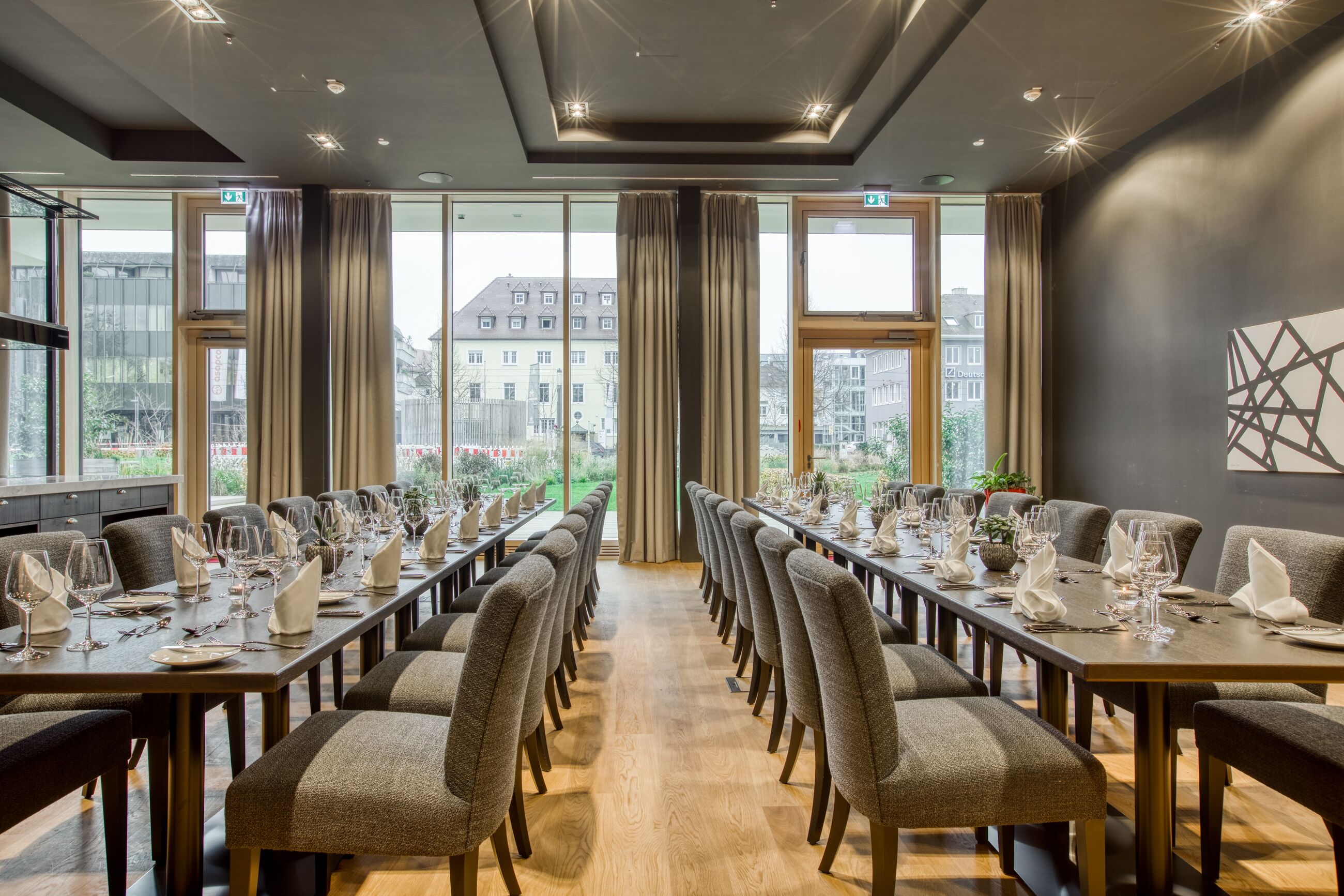 A conference room with long tables set for dining, large windows with curtains, and a view of buildings outside.