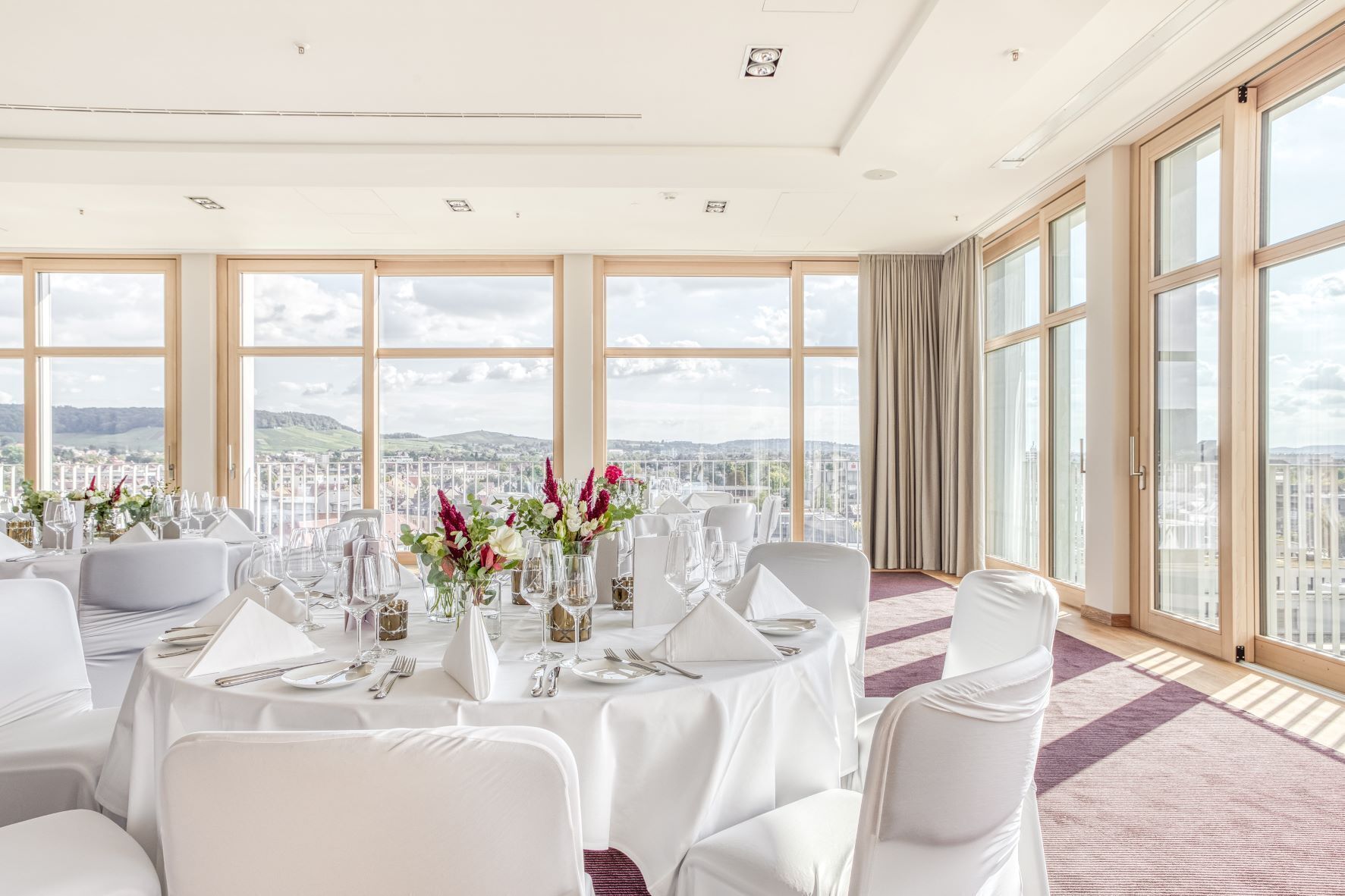 A dining area with round tables set for a meal, large windows offering a city view, and a high ceiling with recessed lighting.