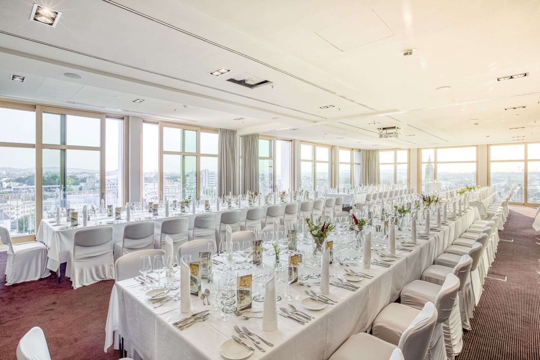 A conference room with a long table set for a formal event, large windows with a city view, and a high ceiling with recessed lighting.