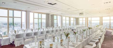 A conference room with a long table set for a formal event, large windows with a city view, and a high ceiling with recessed lighting.