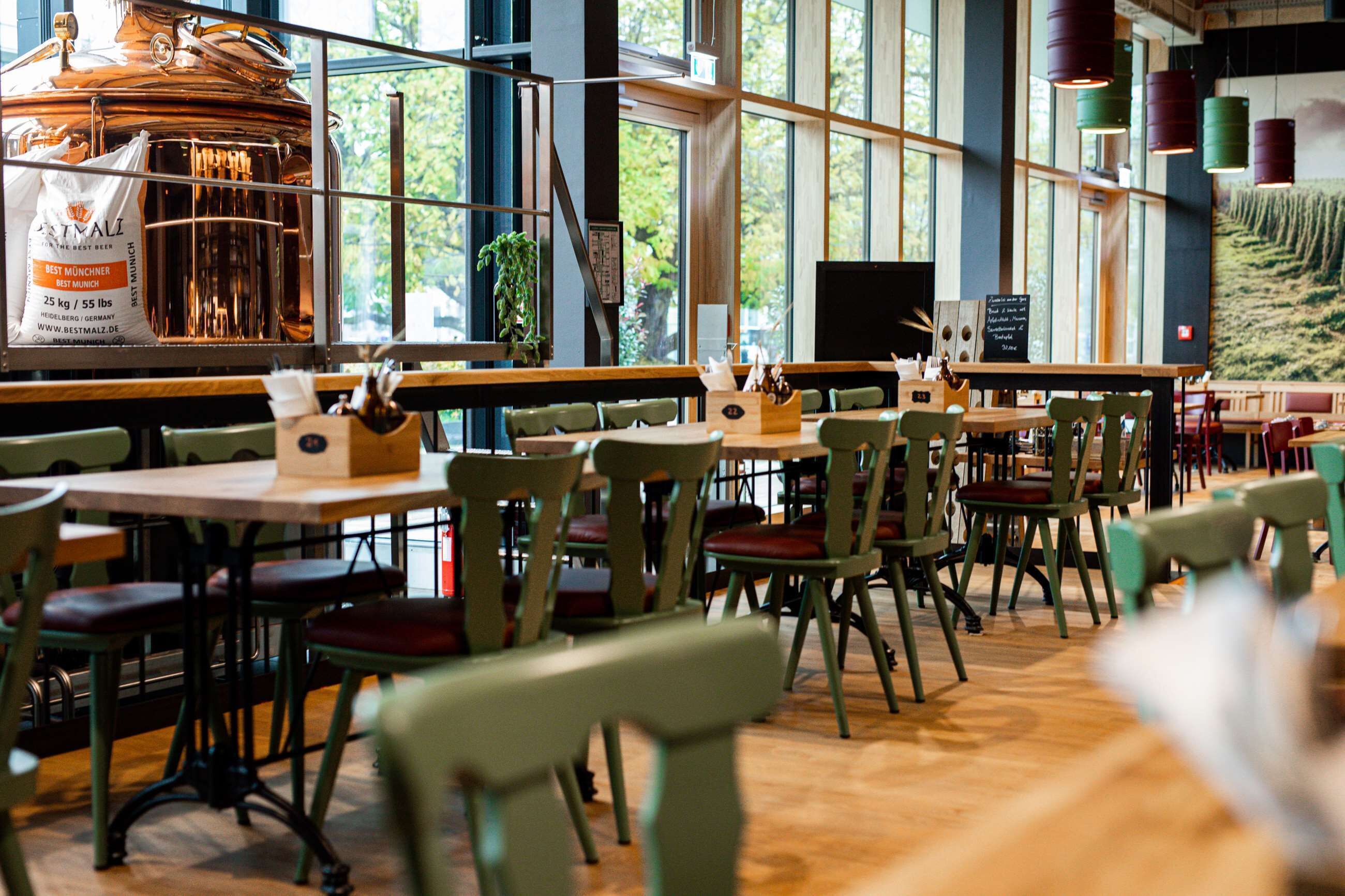 A restaurant interior with wooden tables and chairs, a large copper brewing apparatus, and large windows allowing natural light.