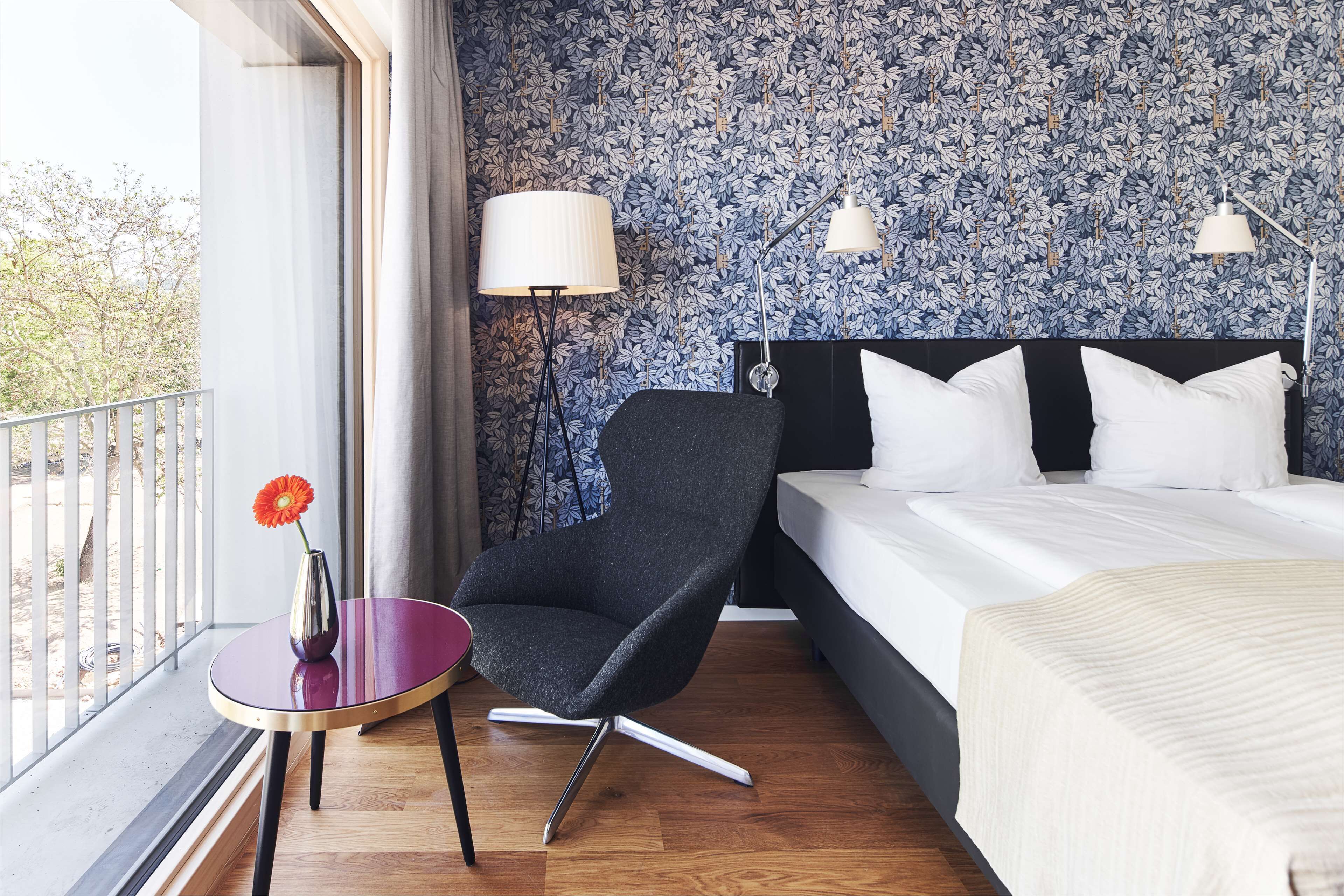 Habitación de hotel con cama, una silla, una mesita con un florero y ventana con vista a árboles.