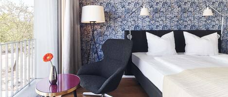 Habitación de hotel con cama, una silla, una mesita con un florero y ventana con vista a árboles.