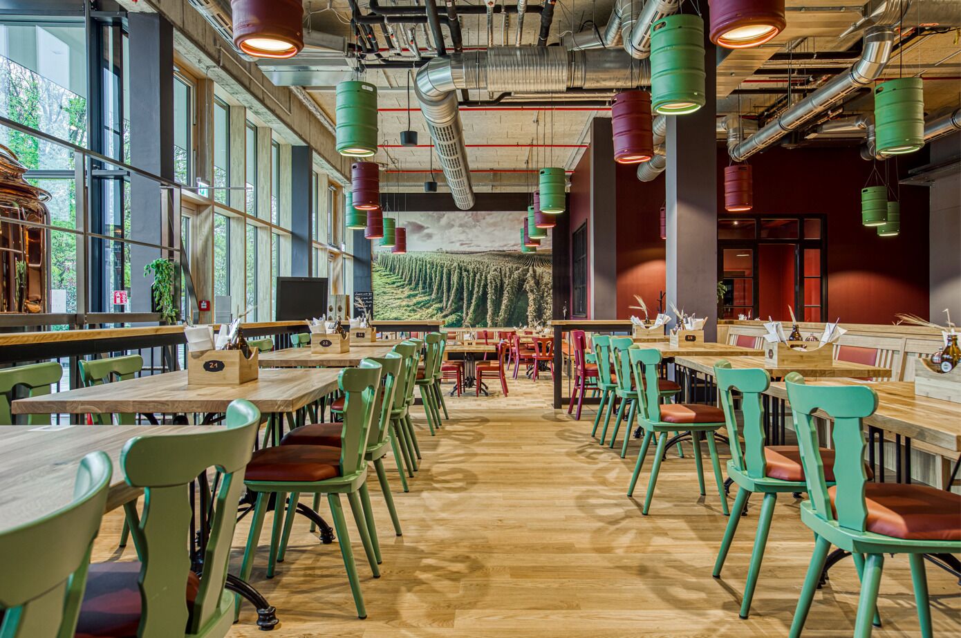 A spacious restaurant with wooden tables and chairs, large windows, and a mural on the wall.