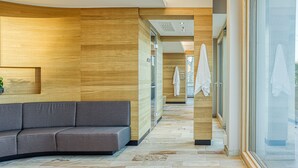 Un hall d’hôtel moderne avec un canapé gris incurvé, des murs recouverts de panneaux en bois et une porte en verre donnant sur une salle de bain où des serviettes sont accrochées au mur.