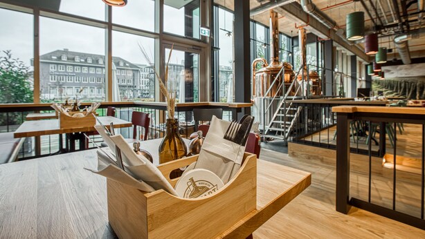 L'intérieur d'un restaurant moderne, avec des tables en bois, un présentoir en bois contenant les couverts et de grandes fenêtres offrant une vue sur la ville.