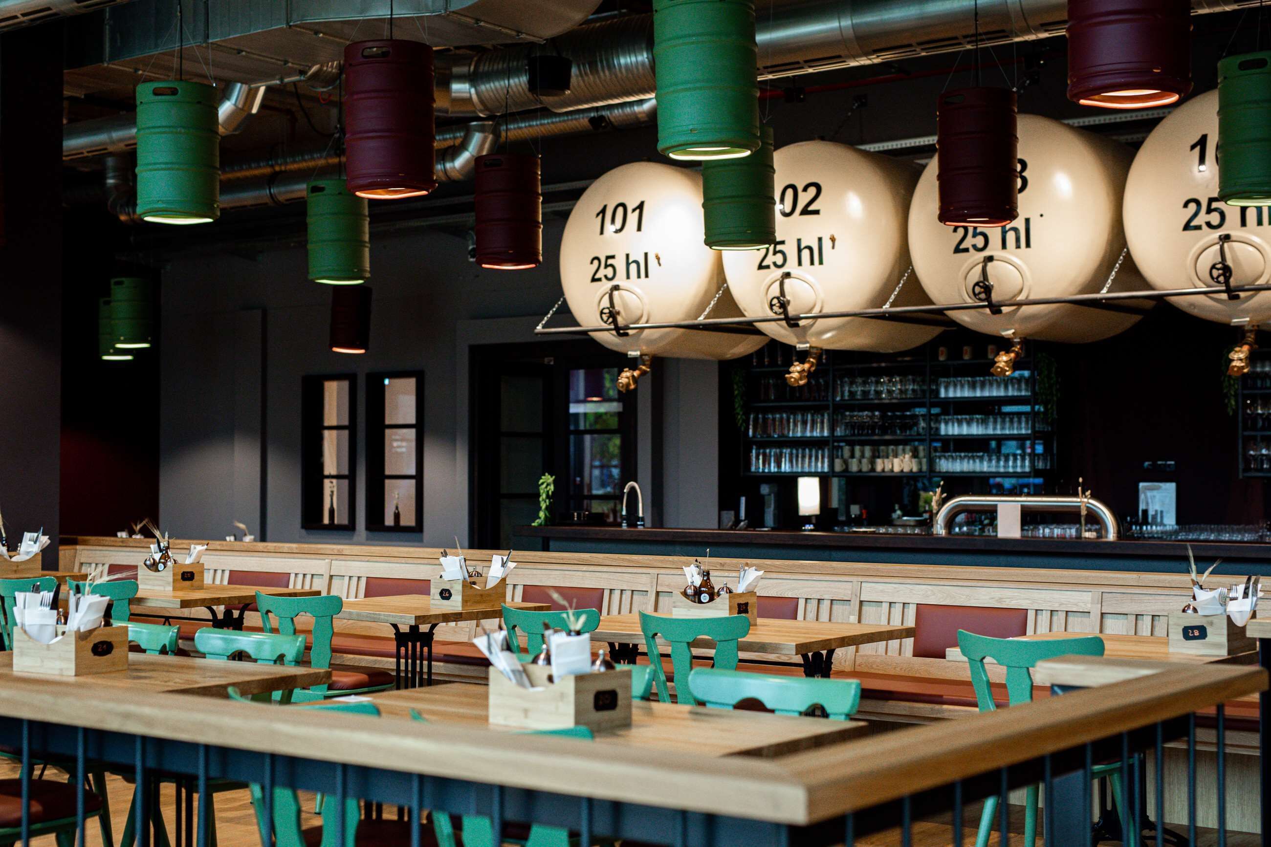 A restaurant interior with wooden tables and green chairs, hanging light fixtures, and a bar area in the background.