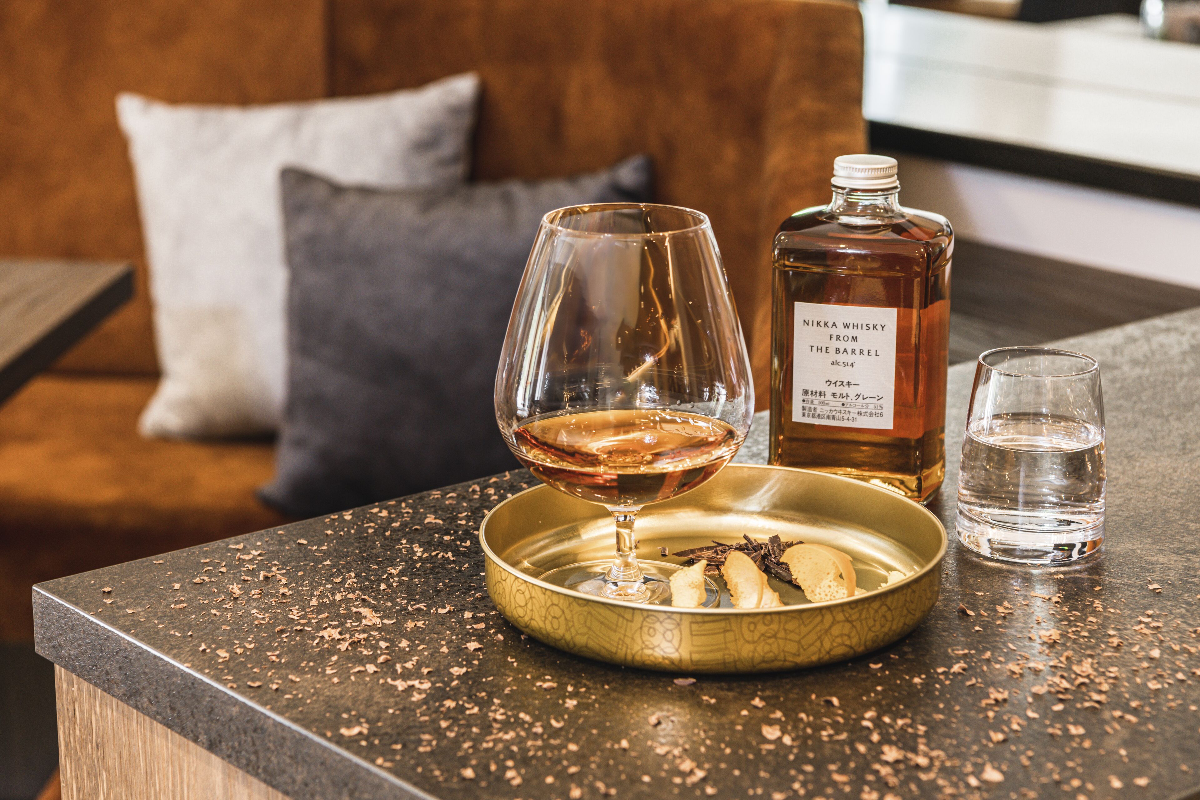 Eine Bar mit einer Whiskeyflasche, einem Whiskeyglas und einem Wasserglas auf einer fleckigen Arbeitsplatte.