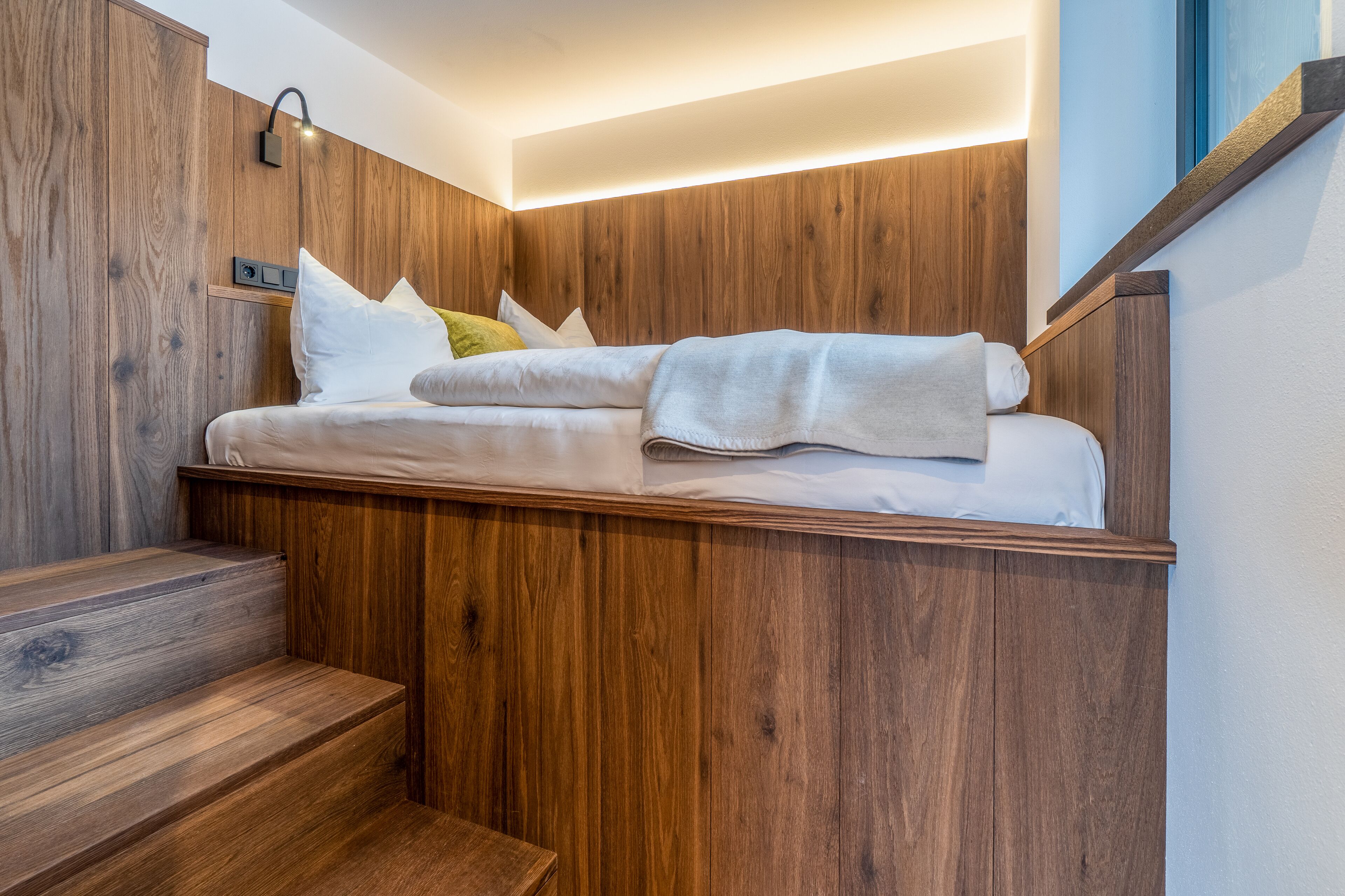 Un dormitorio con una cama frente a una pared de madera, una mesita de noche con una lámpara y una escalera que sube a otro nivel.
