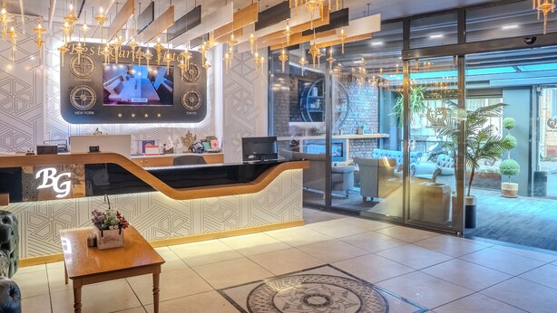 Zona de recepción de un hotel con diseño moderno, que presenta un mostrador curvo, una gran pantalla mostrando un paisaje urbano y un área de descanso con un tapete estampado.